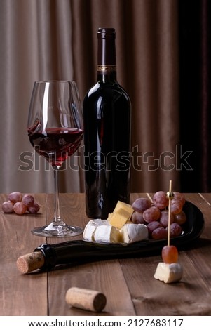 A bottle of red wine, a glass with red wine, a board with snacks, charcuterie board, cheese and grapes on a wooden table, against a background of brown textile
