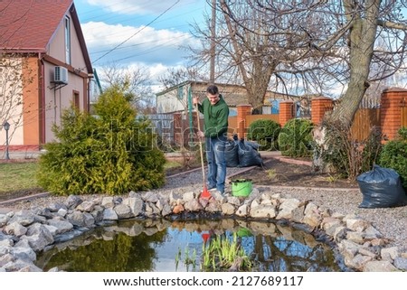 Mature smiling caucasian man cleans a garden pond from water plants and falling leaves. Spring seasonal pond care after winter.