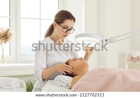 Female cosmetologist performs facial examination procedure at cosmetology clinic. Beautician fixes lamp over face of female patient lying on examination couch in beauty clinic. Cosmetology concept. Royalty-Free Stock Photo #2127702311