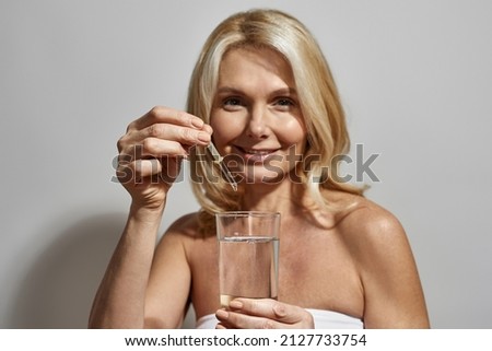 Middle aged caucasian woman dripping medication from pipette in glass with water. Smiling blonde female person looking at camera. Health treatment and care. Isolated on white background in studio