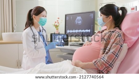 asian pregnant woman wearing protective face mask to prevent COVID19 lying in hospital ward has uterus utltrasonographic diagnosis - female doctor shows picture on computer and digital tablet