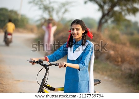 Portrait of happy young rural indian girl wearing blue school uniform standing with bicycle at village street. Royalty-Free Stock Photo #2127759794