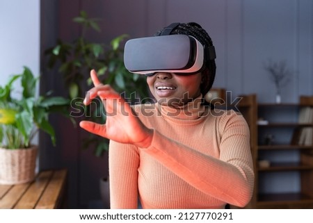 Young woman enjoy vr gaming, using virtual reality interface for shopping wearing goggles at home. African lady excited first virtual reality experience try to touch objects in digital simulation app Royalty-Free Stock Photo #2127770291