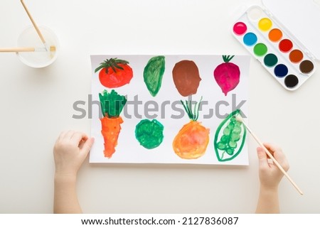 Baby hand holding paint brush and drawing different colorful vegetable shapes on white paper on light table background. Closeup. Point of view shot. Toddler development.