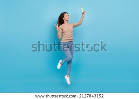 Photo of cheerful lady jump hold phone take selfie show v-sign wear beige sweater isolated blue color background