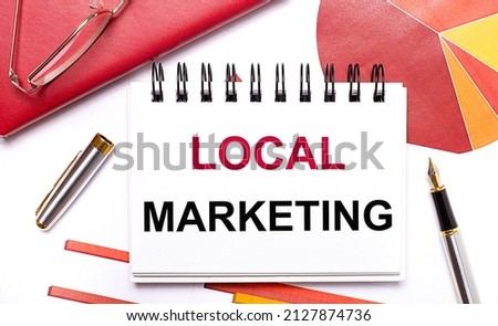 On the desktop is a white notebook with the text LOCAL MARKETING, a pen, burgundy and red tables, and gold-framed glasses. Business concept.