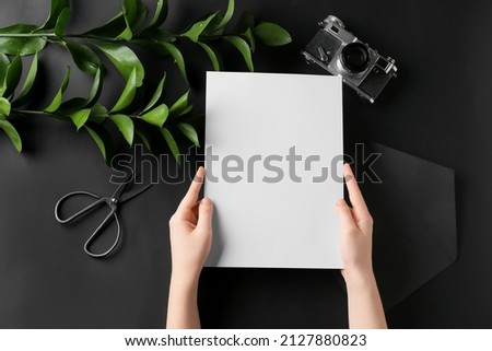 Female hands with blank sheet of paper, scissors, plant branches and photo camera on dark background
