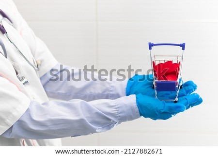 The hands of a doctor in a white medical gown and rubber gloves with a phonendoscope hold a grocery cart with red hearts in a hospital. Selective focus. Close-up