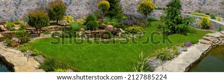 Lush green botanical garden - blooming spring flowers and lawn path,with grass growing up between the stones.Beautiful small garden pond with stone banks and ornamental plants. Royalty-Free Stock Photo #2127885224
