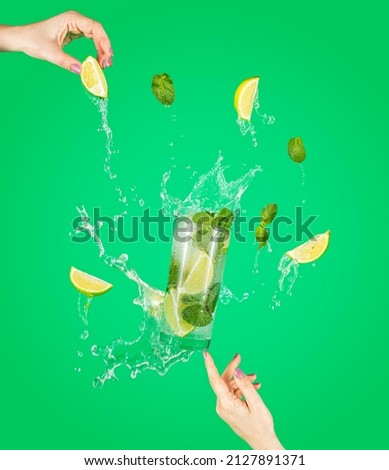 Woman hand support fly glass of tasty Mojito cocktail with lime and mint with splash, ingredients falling to glassful. Summer art food concept on green background