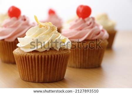 Picture of a cupcake with a yellow butter cream frosting, sprinkled with light  golden sugar and decorated with lemon zest on the top. Other cupcakes blurry visible in the background.
