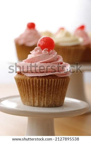 Close-up picture of a cupcake with a pink butter cream frosting, decorated with a cocktail cherry on a top. Parts of other cupcakes blurry visible in the background.