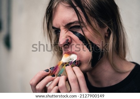 Tight headshot of an attractive emo punk young man with long hair devouring sweet gummy bears. Close up of a pierced and face painted guy in  his late 20s with mouth full of jelly candies. 