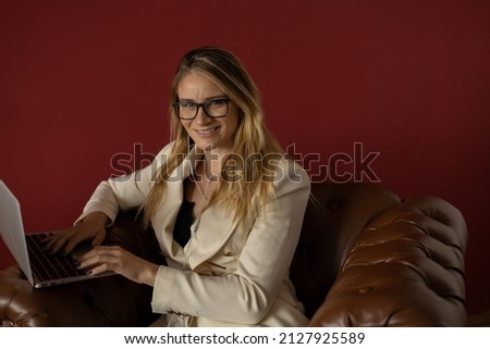 Young woman working at home with laptop and papers on a classic brown arm chair and headphones.  Red painted wall background. Home office concept. Gray notebook for working. High quality photo