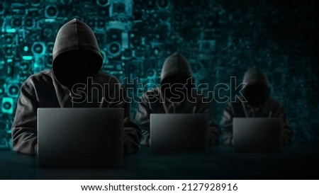 Hacker with a hidden face and with a laptop sits at the table, blue background. There is an hourglass on the hacker's desk. Place for text. High quality photo Royalty-Free Stock Photo #2127928916