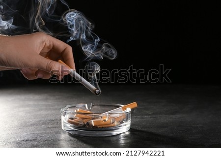 Woman holding smoldering cigarette over glass ashtray at grey table against black background, closeup Royalty-Free Stock Photo #2127942221