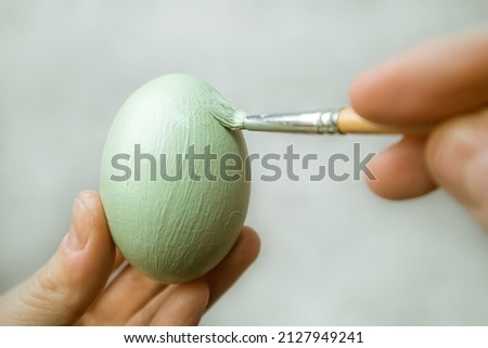 Hand holds brash and paints egg. A process of painting an Easter egg with light green paint. Paint brush in woman's hand on gray background. Tradition and people concept. Spring season, April holiday. Royalty-Free Stock Photo #2127949241