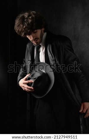 Young man with beard in white shirt and black leather coat with hat against black background.