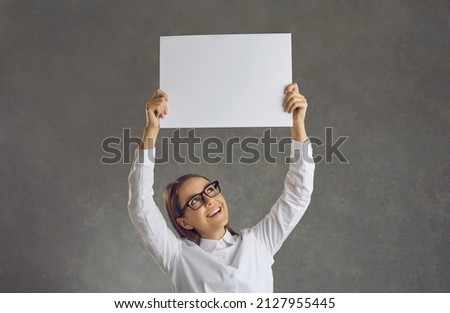 Happy smiling Caucasian woman looks at a blank sheet of paper she holds over her head. Woman makes an announcement, votes or expresses her position, standing on a gray background. Promotion concept.