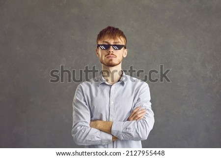 Haughty bearded film fan with pixel glasses, person with cross his arms, portrait, gray background. Sly young guy. Man stands, arms crossed. Cool man with glasses.