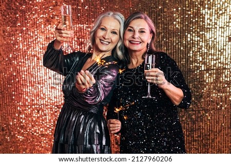 Two happy senior women holding sparklers and glasses of champagne against a glitter background