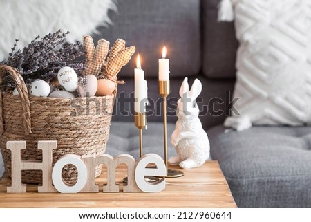 Provence. A wicker basket with Easter eggs, lavender, candles and white rabbits in the interior of the living room on a wooden table. The concept of home comfort in the bright holiday of Easter 2022. Royalty-Free Stock Photo #2127960644