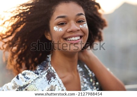 Close up portrait of overjoyed charming african american woman with curly hair and glitter on cheekbones smiling happily at camera while moving dancing outdoors, festive atmosphere in the air Royalty-Free Stock Photo #2127988454