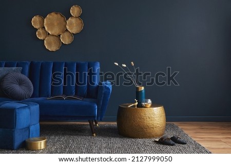 Stylish living room interior composition with velvet blue sofa, golden side table, pouf, pillows and elegant home decor. Dark blue wallpaper. Template. Copy space. Royalty-Free Stock Photo #2127999050
