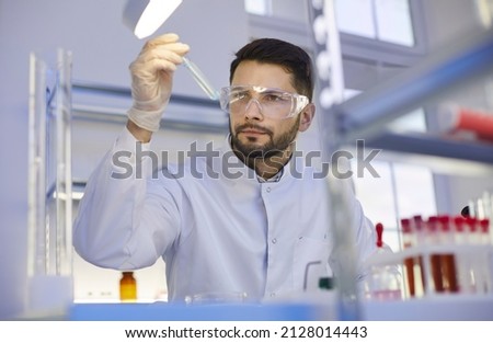 Young Caucasian man in white medical uniform and glasses do laboratory experiments with test tube. Male assistant do chemistry investigation research in lab. Biotechnology, science concept. Royalty-Free Stock Photo #2128014443