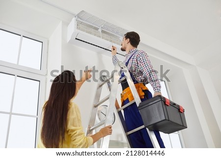 Technician from the AC Maintenance and Repair Service climbs a ladder with his toolbox in order to check or fix troubles with a modern white wall mounted air conditioner in a young lady's home Royalty-Free Stock Photo #2128014446