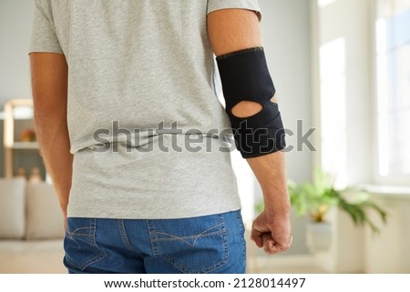 Young man wearing an adjustable black neoprene orthopedic elbow support brace on his right arm for easing pain in the elbow, back view, cropped close up shot. Concept of physical injury treatment Royalty-Free Stock Photo #2128014497