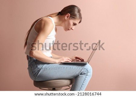 Woman with bad posture using laptop while sitting on stool against pale pink background Royalty-Free Stock Photo #2128016744
