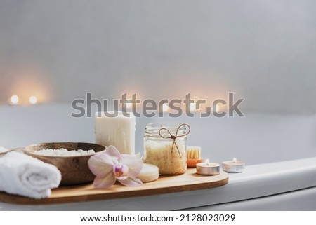 Preparation for hotel spa treatment, home bath procedure. White washbasin in bathroom, accessories on tray. Burning candles, soap, foot brush, towel, glass bottle with sea salt, orchid flower Royalty-Free Stock Photo #2128023029