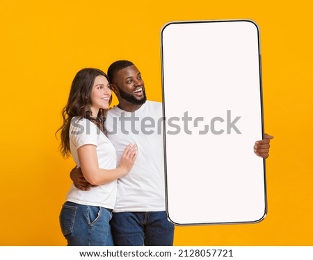 Great Offer. Portrait of smiling interracial couple hugging and holding big giant cell phone looking at white blank device screen in hand, presenting gadget with free copy space for mock up, ad banner Royalty-Free Stock Photo #2128057721
