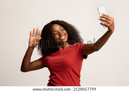 Happy Young Black Woman With Smartphone Making Video Call Or Taking Selfie, Portrait Of Cheerful African American Female Waving Hand At Mobile Phone Camera While Standing Over White Studio Background Royalty-Free Stock Photo #2128057799