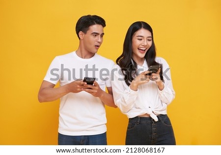Shocked asian man spying on her smiling girlfriend while both using mobile phones isolated over yellow background. chatting or social media concept. Royalty-Free Stock Photo #2128068167