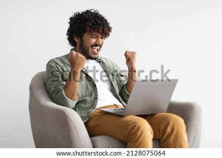 Emotional curly bearded young hindu guy in casual sitting in armchair with modern laptop and clenching fists, celebrating success, trading on stocks and markets online, got good profit, copy space Royalty-Free Stock Photo #2128075064