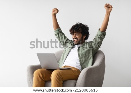 Happy young indian man in casual sitting in armchair with computer on his lap, looking at laptop screen and raising hands up, unemployed guy got new job, copy space. Job search websites concept Royalty-Free Stock Photo #2128085615