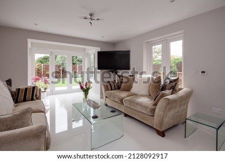 Stunning living room with glass coffee tables, gloss white porcelain tiled floor, white walls, sofas with decorative cushions and bifold patio door leading to garden Royalty-Free Stock Photo #2128092917