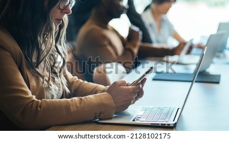 An Anonymous Businesswoman Using Mobile Phone in Open Plan Office. 
Unrecognizable team of business people sitting in a row at same table and working together on a laptop computers and smartphones. Royalty-Free Stock Photo #2128102175