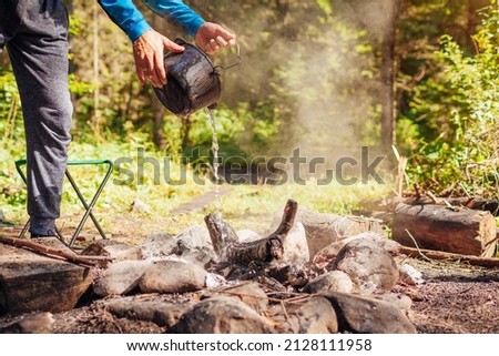 Man extinguishing campfire with water from cauldron in summer forest. Put out campfire by tent. Traveling fire safety rules Royalty-Free Stock Photo #2128111958