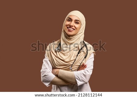 Portrait of a female Muslim doctor wearing a white coat, arms crossed, standing against a brown background and smiling. The concept of medicine, care, health. Royalty-Free Stock Photo #2128112144