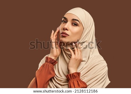 Portrait of beautiful modern muslim woman with natural make-up dressed in beige hijab posing on brown background in studio. Facial skin care, female beauty. Royalty-Free Stock Photo #2128112186