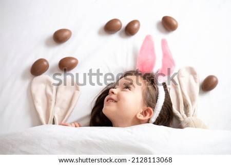 Happy Easter. cute beautiful girl with blue eyes and bunny ears peeking out from under the blanket. Chocolate eggs are scattered on the bed. Egg hunt. Lifestyle High quality photo