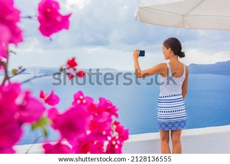 Travel tourist woman taking pictures with phone of Santorini Mediterranean sea view from coast of greek island. Europe Greece destination girl sightseeing Oia in spring with pink flowers foreground.