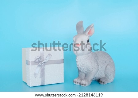 Rabbit sculpture on a blue background and a box with a silver ribbon as a symbol of Easter holidays and gifts. Copyspace. Place for text. Congratulatory banner, postcard for easter.