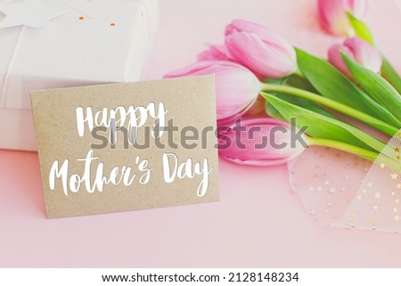 Happy mothers day text on greeting card, pink tulips bouquet and gift box on pink background. Stylish greeting card. Happy Mother's Day, gratitude and love to mom. Handwritten lettering