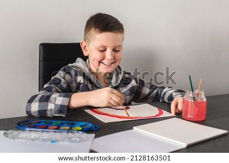 Smiling boy with dimples having fun and drawing picture with colorful paints and brushes. Kid drawing with paintbrushes and watercolors. Child painting rainbow picture, creativity at home concept.