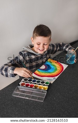 Smiling boy with dimples having fun and enjoy painting rainbow in his album. Kid drawing with paintbrushes and watercolor paints. Schoolboy child painting colorful rainbow picture sitting at home.