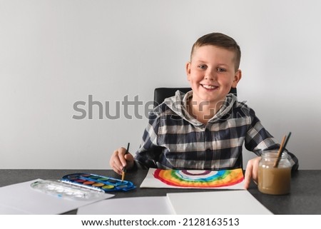 Boy having fun and enjoy painting rainbow in his album. Smiling kid with dimples drawing with paintbrushes and watercolor paints. Caucasian child painting rainbow picture, creativity at home concept.